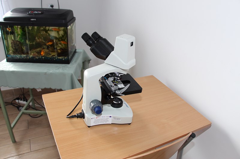 Obrazek przedstawia zdjęcie mikroskopu stojącego na biurku, i niewielki akwarium na innym biurku za nim