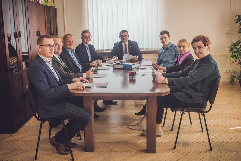 Obrazek przedstawia 8 ludzi siedzących za jednym biurkiem 