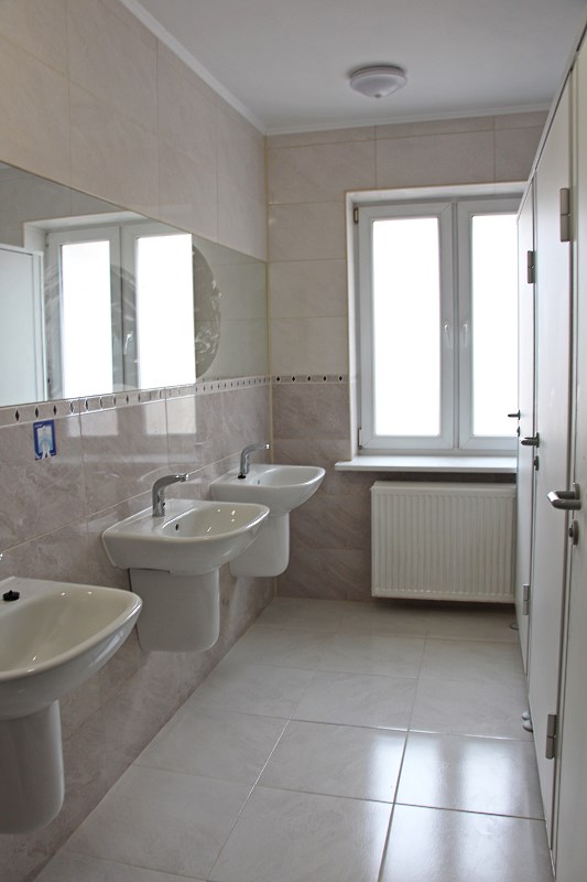 Obrazek przedstawia zdjęcie łazienki - są widoczne 3 umywalki i wielkie lustro na ścianie nad nimi, okno i bateria grzewcza