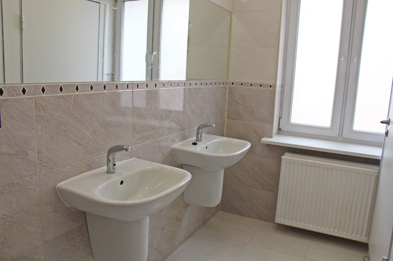 Obrazek przedstawia zdjęcie z łazienki- są widoczne dwie umywalki z wielkim lustrem na ścianie nad nimi, na innej ścianie - okno i bateria pod nim
