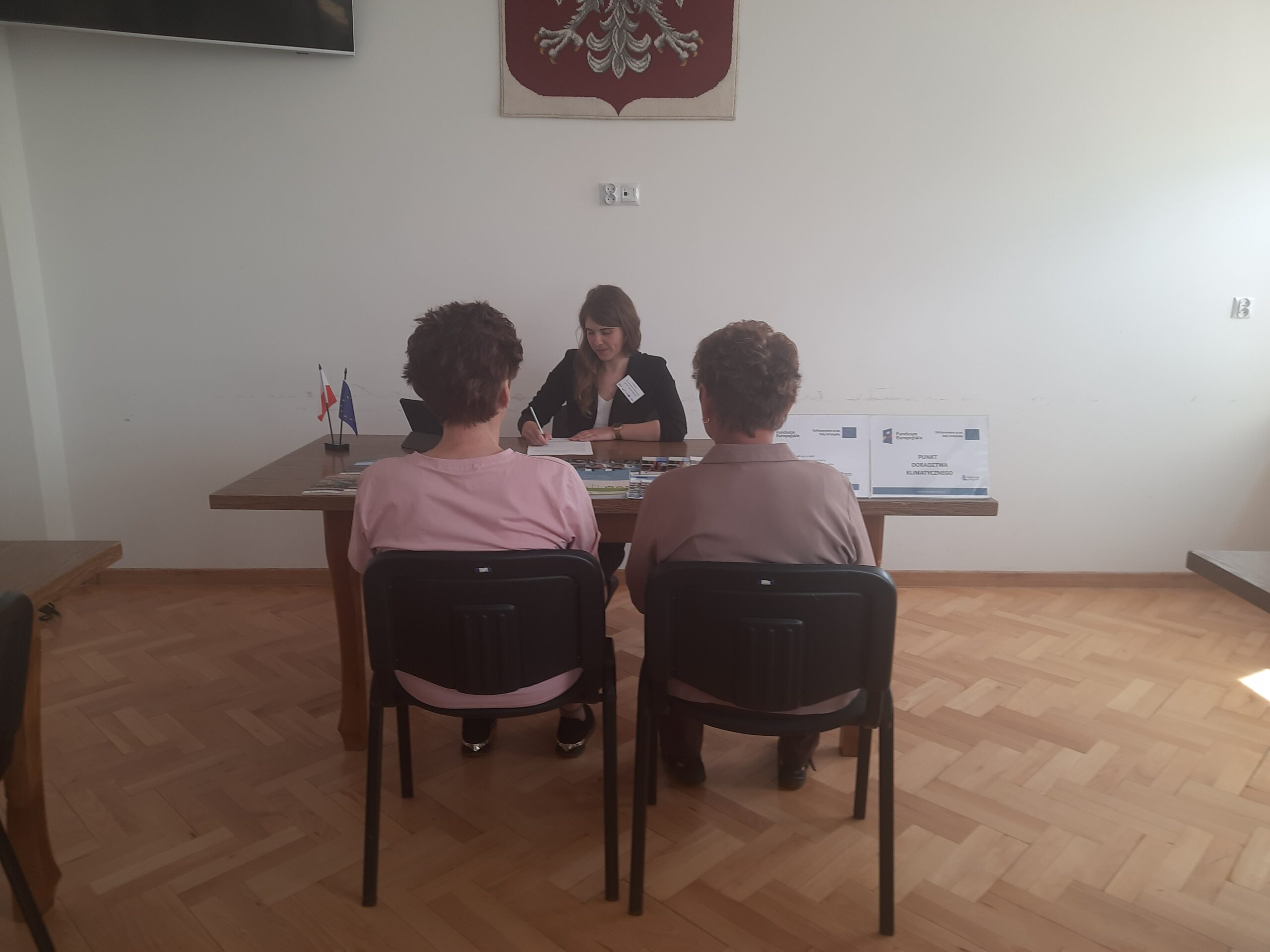 Doradca klimatyczny wraz z interesantami udzielający bezpłatnych konsultacji w zakresie neutralności klimatycznej wraz z uzupełnianiem kart doradztwa w sali narad podczas realizacji Mobilnego Punktu Doradztwa Klimatycznego w Urzędzie Gminy Chrzanów w dniu 28 maj 2025 r. w oddali