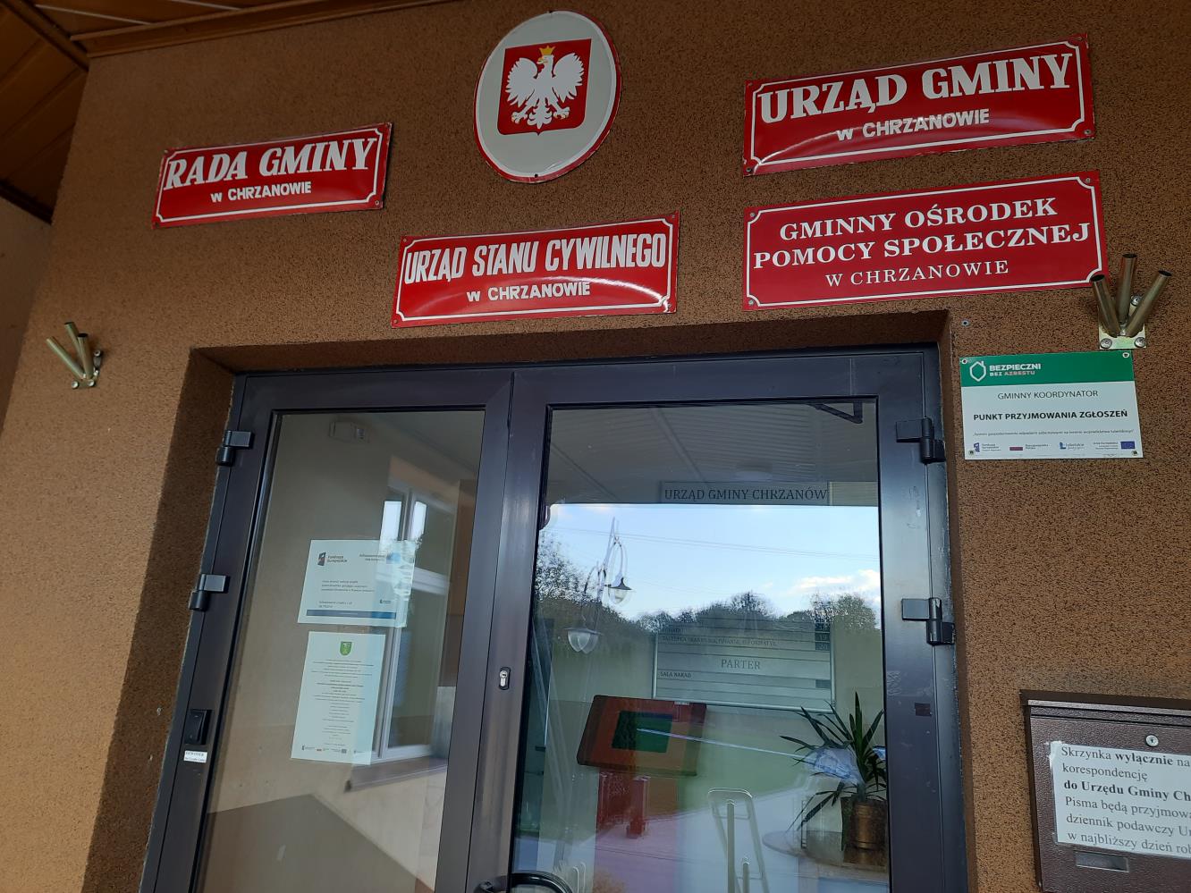 Plakat – Powiat Janowski realizuje projekt: System doradztwa sprzyjający osiągnięciu neutralności klimatycznej w Powiecie Janowskim wraz z zaproszeniem na bezpłatne konsultacje wywieszony na drzwiach wejściowych budynku Urzędu Gminy Chrzanów