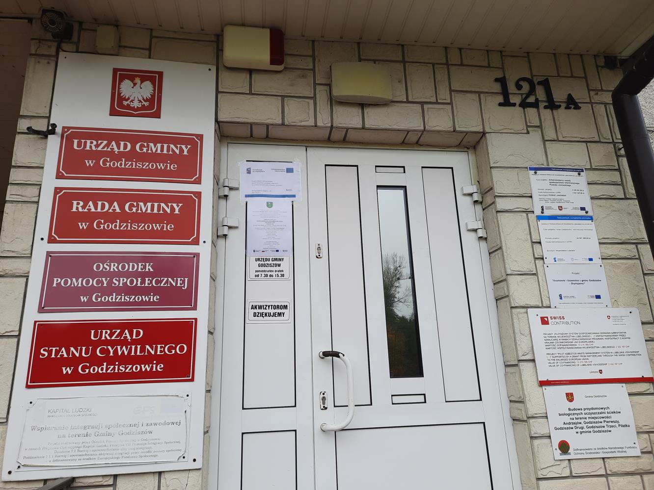 Plakat – Powiat Janowski realizuje projekt: System doradztwa sprzyjający osiągnięciu neutralności klimatycznej w Powiecie Janowskim wraz z zaproszeniem na bezpłatne konsultacje wywieszony na drzwiach wejściowych budynku Urzędu Gminy Godziszów