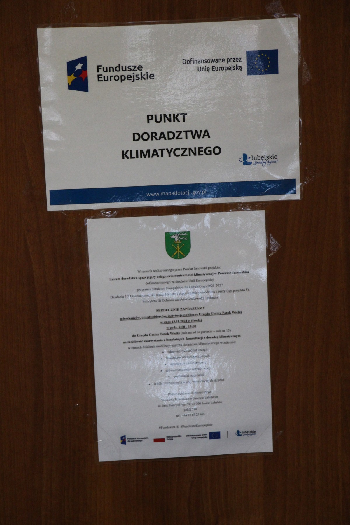 Plakat – Powiat Janowski realizuje projekt: System doradztwa sprzyjający osiągnięciu neutralności klimatycznej w Powiecie Janowskim wraz z zaproszeniem na bezpłatne konsultacje wywieszony na drzwiach Sali narad w Urzędzie Gminy Potok Wielki