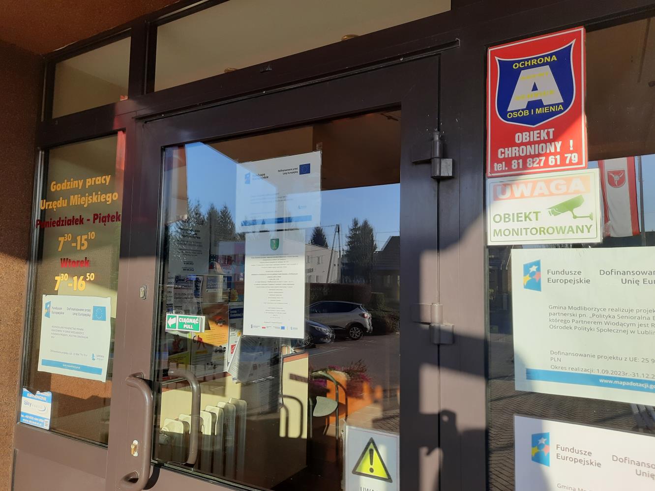 Plakat – Powiat Janowski realizuje projekt: System doradztwa sprzyjający osiągnięciu neutralności klimatycznej w Powiecie Janowskim wraz z zaproszeniem na bezpłatne konsultacje wywieszony na drzwiach wejściowych budynku Urzędu Miejskiego w Modliborzycach