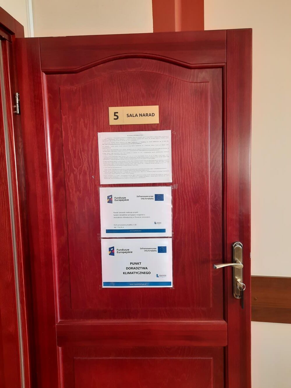 Plakat – Powiat Janowski realizuje projekt: System doradztwa sprzyjający osiągnięciu neutralności klimatycznej w Powiecie Janowskim wraz z plakatem Doradztwa Klimatycznego wywieszony na drzwiach sali narad Urzędu Miejskiego w Modliborzycach