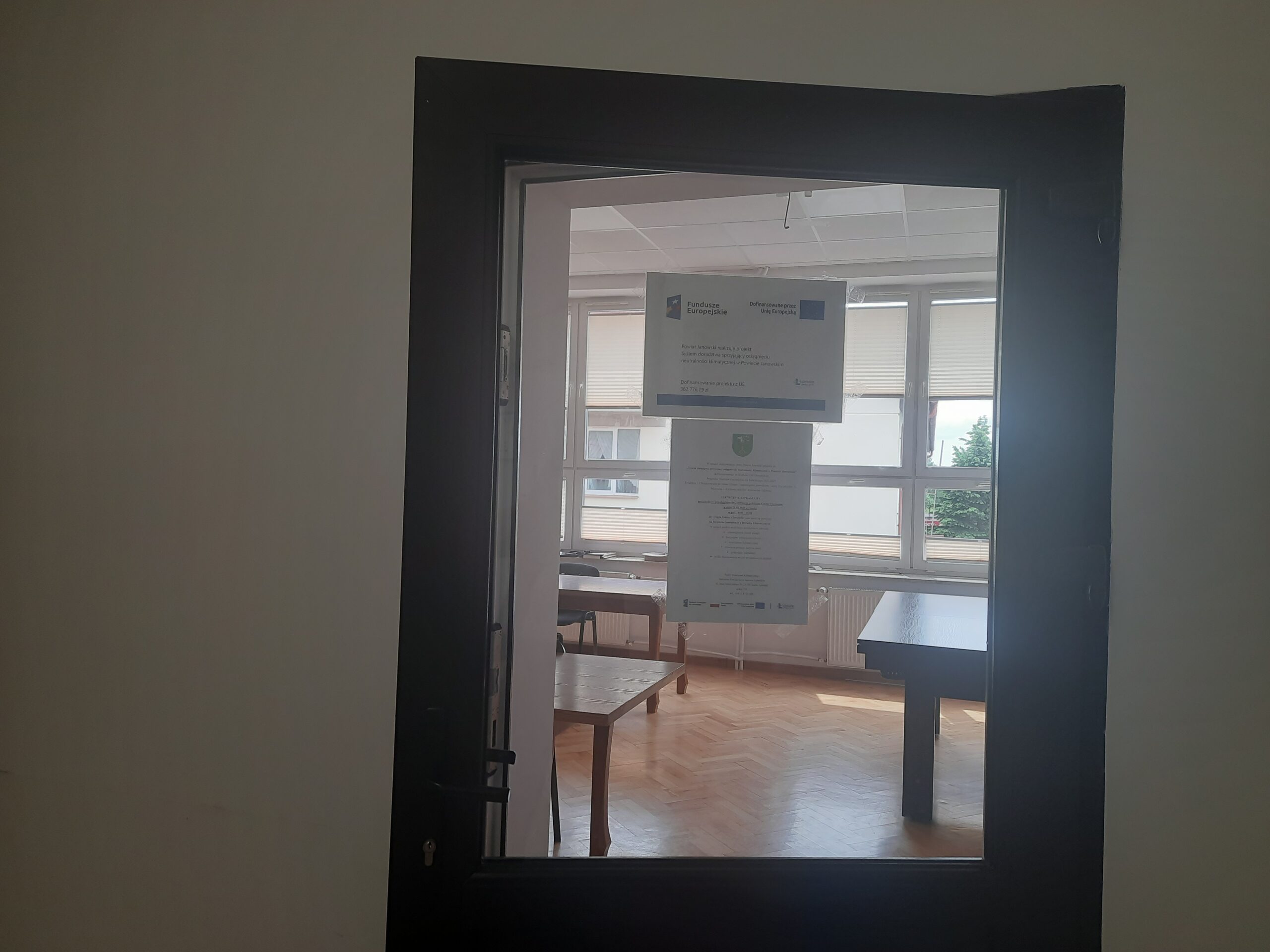 Plakat - Powiat Janowski realizuje projekt: System doradztwa sprzyjający osiągnięciu neutralności klimatycznej w Powiecie Janowskim wraz zaproszeniem na bezpłatne konsultacje wywieszony na drzwiach sali narad Urzędu Gminy Chrzanów